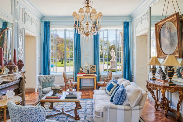 Image of a light and airy room, featuring light blue walls and white ceiling, rugs and chairs that are blue, a painting on the wall, overlooking a grassy meadow.