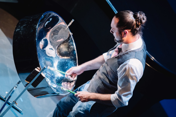 Image of a man. hair styled into a bun, playing the steel pan.