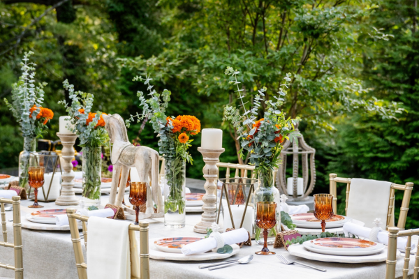 Image of a beautiful table scape featuring orange flowers, eucalyptus, and white linens, set against a backdrop of lush green trees.
