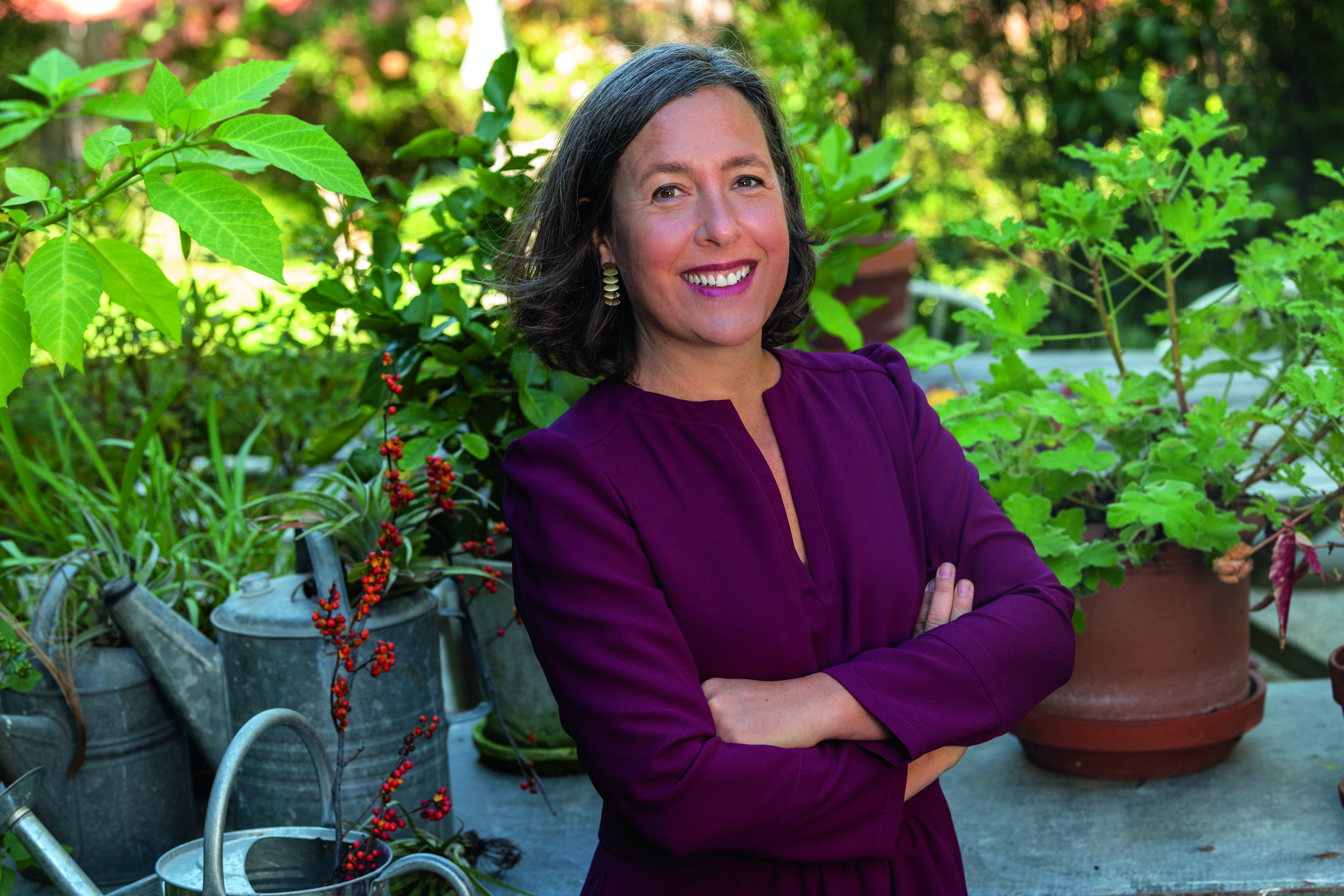 Headshot of author Nicole Juday, posing with her arms crossed and smiling in front of a garden of potted plants.