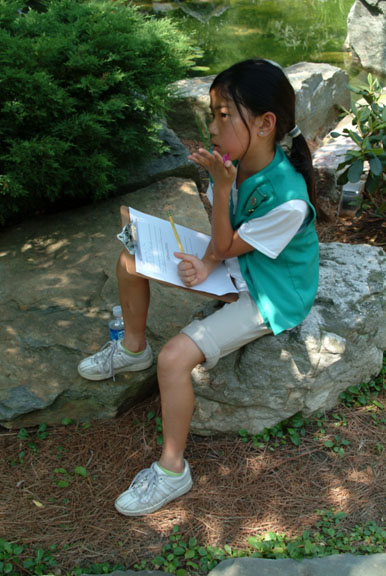 A Junior Girl Scout wearing a green vest sits on a large rock in a garden holding a clipboard and pencil with her chin in her hand staring contemplatively into the distance.