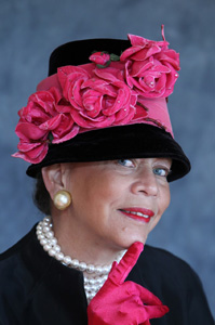 Donna Limerick wears a black dress, pearls, and a black hat with hot pink roses and hot pink gloves. 