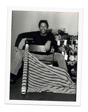 Black and white photo of designer Jay Jaxon sitting backward on a chair, surrounded by bolts of fabric