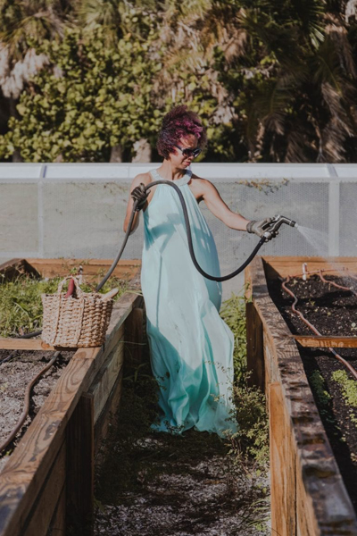 Gesel Mason holds a hose, watering a garden of raised beds in a light blue, chiffon evening gown