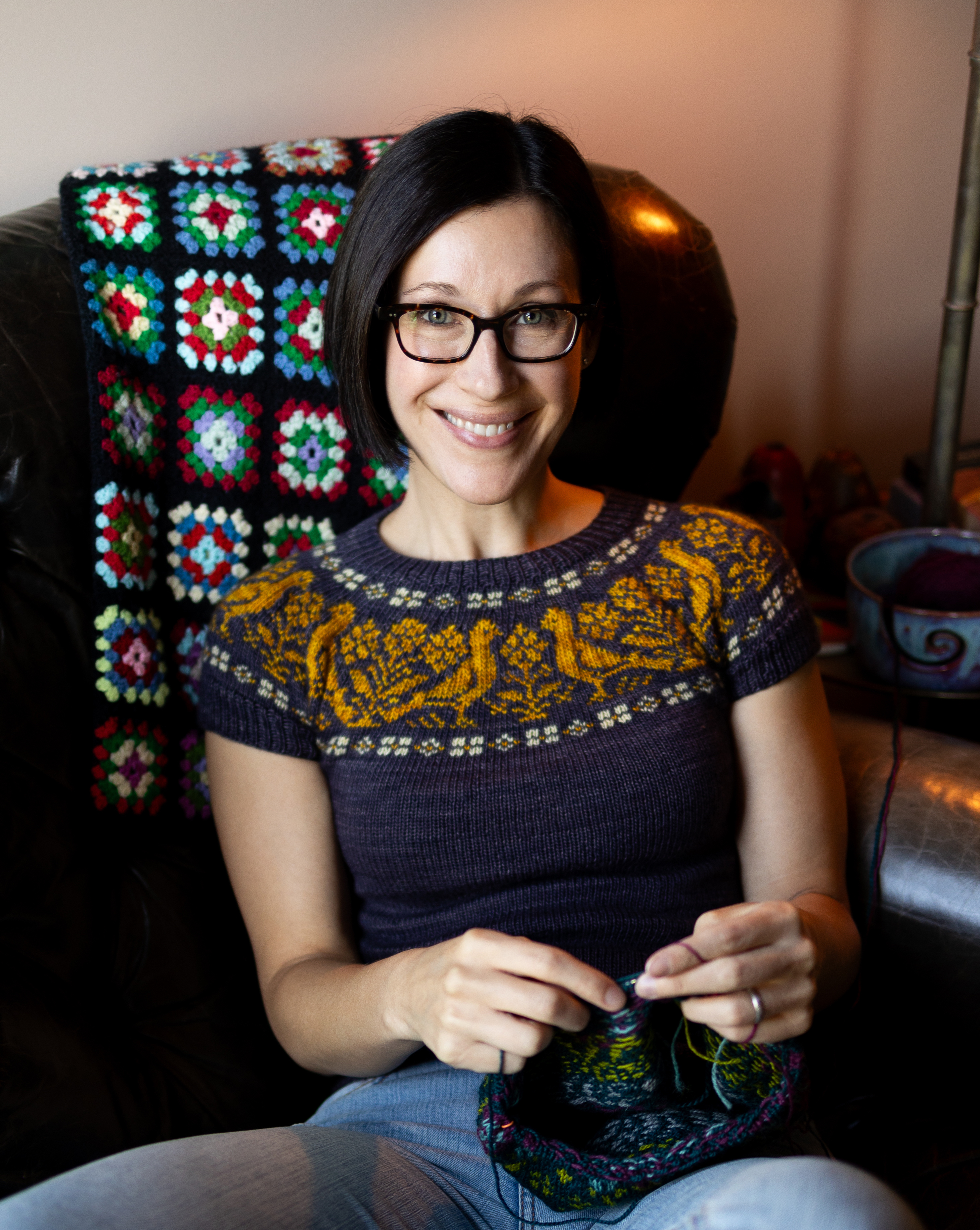 Image of Nicole Nehrig, with short brown hair, glasses, and wearing a knit sweater, sitting in a chair knitting.