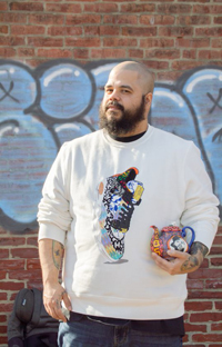 Photograph of Roberto Lugo holding one of his teapots, standing in front of a brick wall with graffiti art