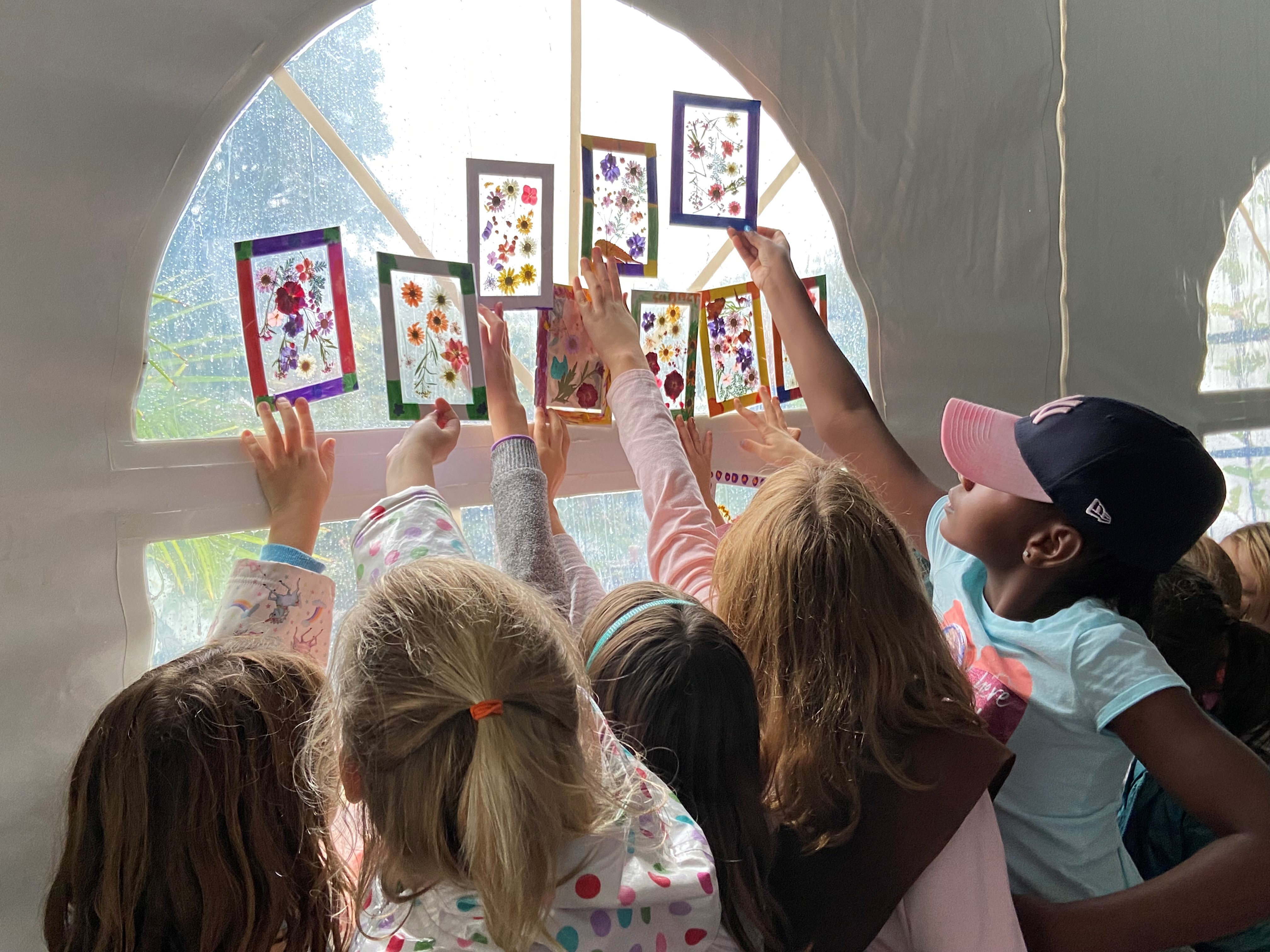 Girl Scouts hold their pressed flower suncatchers up to a window so the sunlight shines through their artwork.