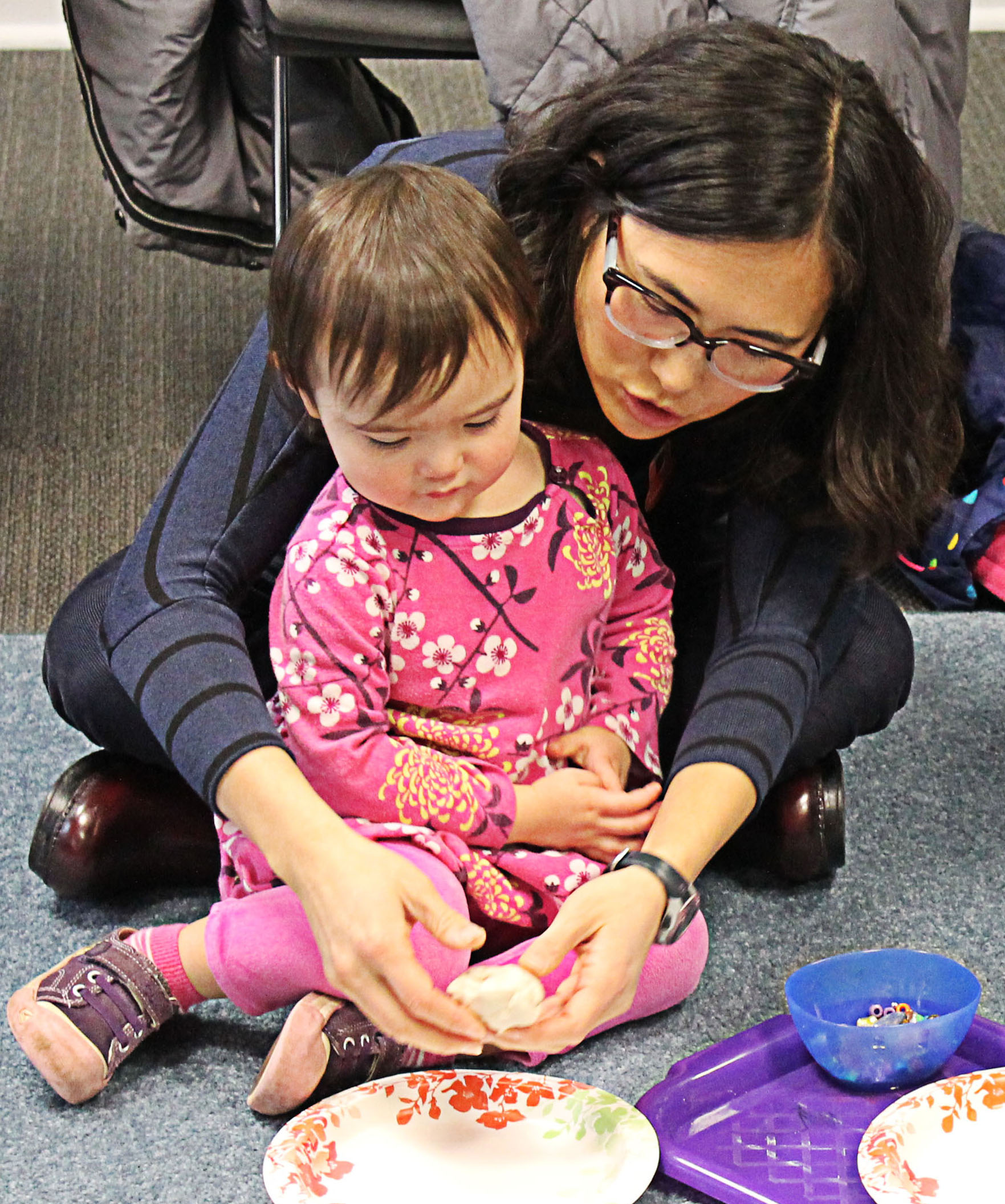 A parent and toddler child sit on a carpet working together on an art project.