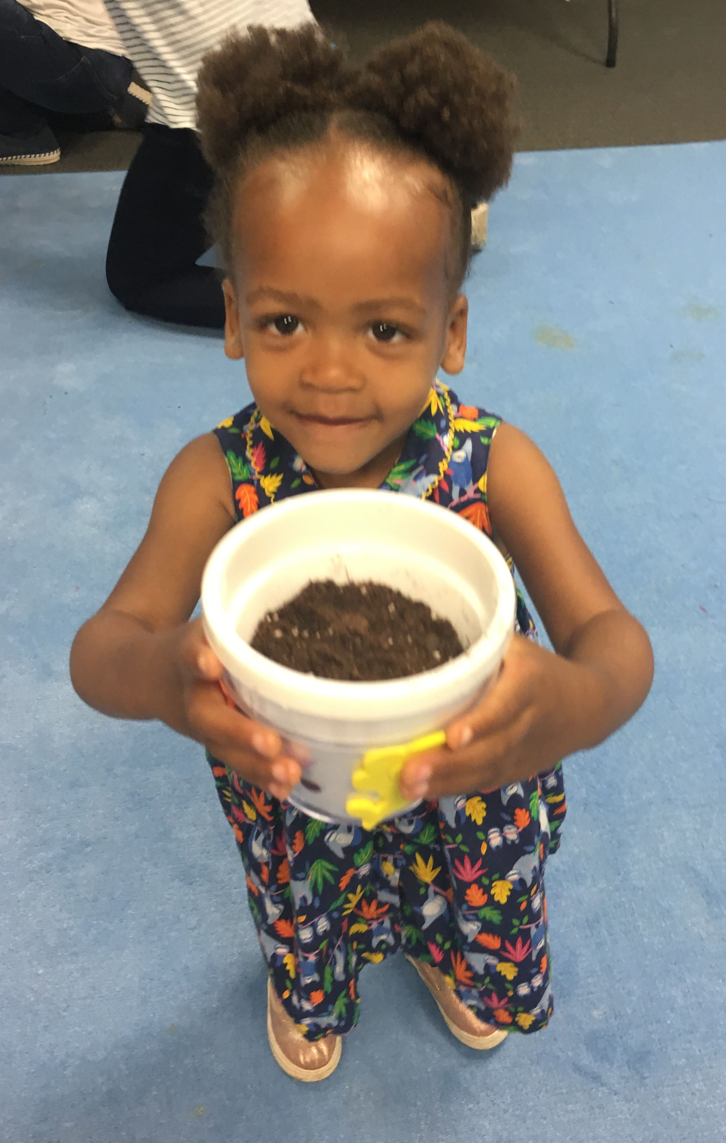 A preschooler holds up her small flower pot filled with soil, ready to plant a seed.