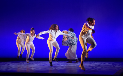 Five dancers dressed in all white performing against a deep blue background