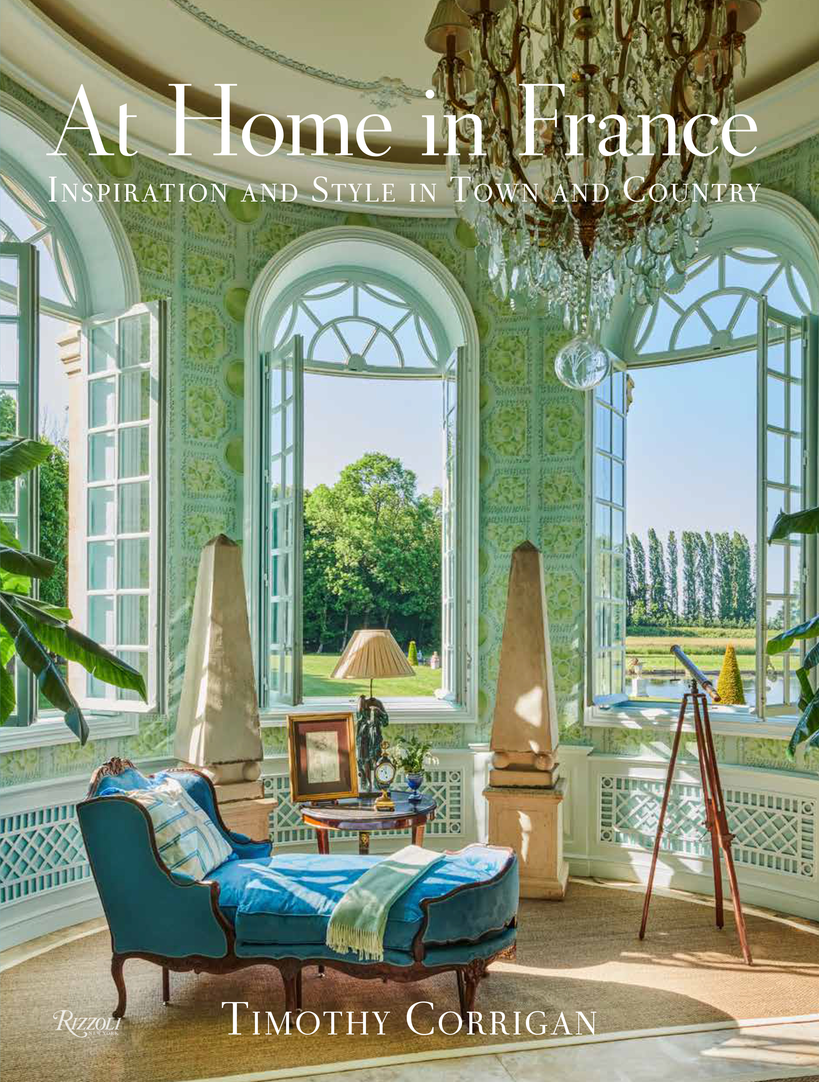 Book cover for "At Home in France" by Timothy Corrigan. The cover features an airy room with a blue lounge chair by a set of open windows overlooking a grassy meadow.