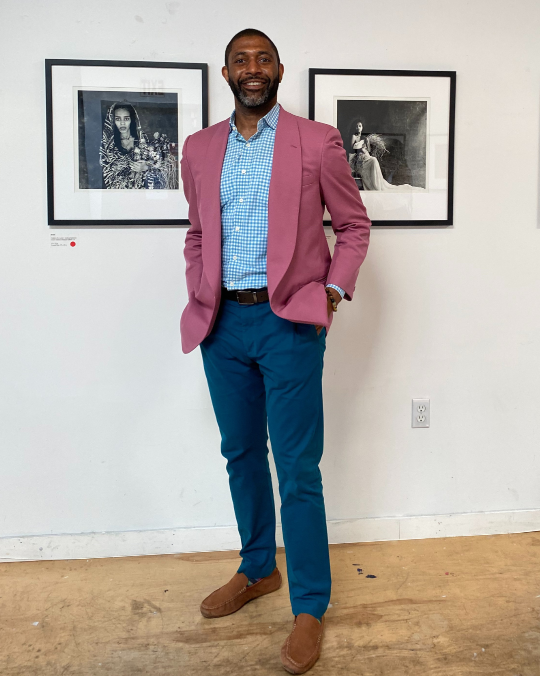 Image of Adrian Ferguson standing in front of artwork hanging on the wall. He is wearing a pink suit jacket and posing with his hands in his pockets. 