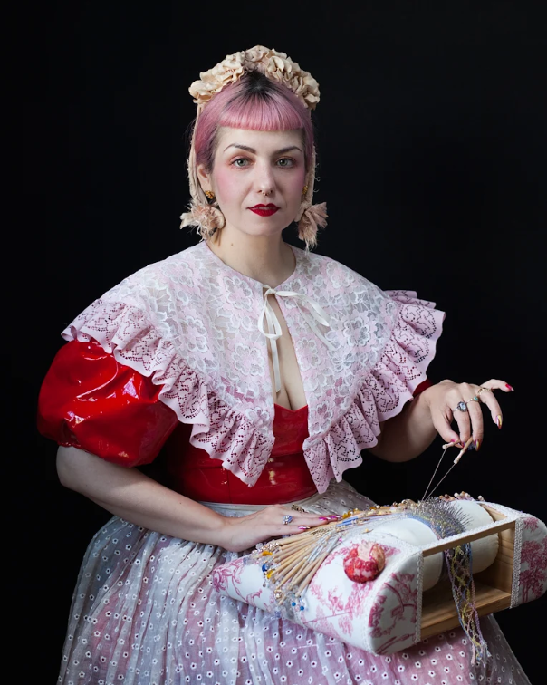 Image of Elena Kanagy-Loux, with pink hair, a flower bonnet, and intricate lace shawl, sitting and creating bobbin lace.