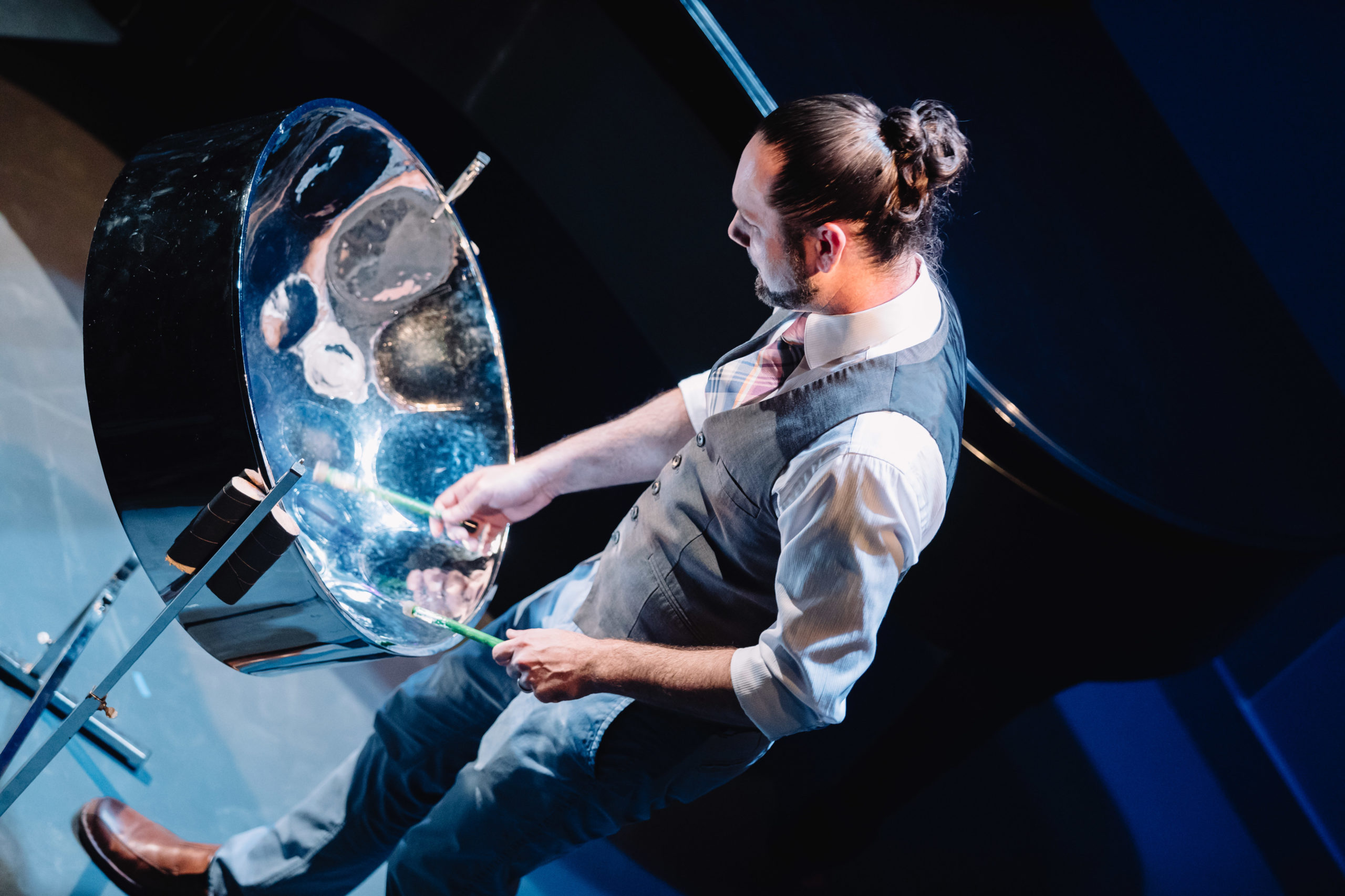 Image of a man, hair styled into a bun, playing the steel pan.