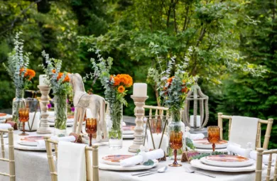 Image of a beautiful table scape featuring orange flowers, eucalyptus, and white linens, set against a backdrop of lush green trees.