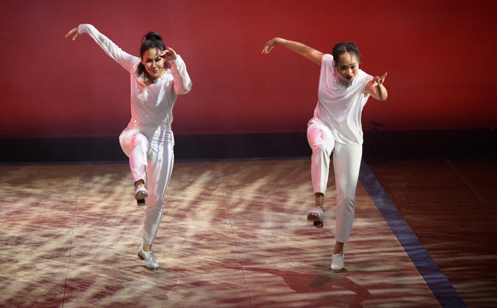 Two dancers wearing all-white, paused as if mid-stomp, against a deep red background