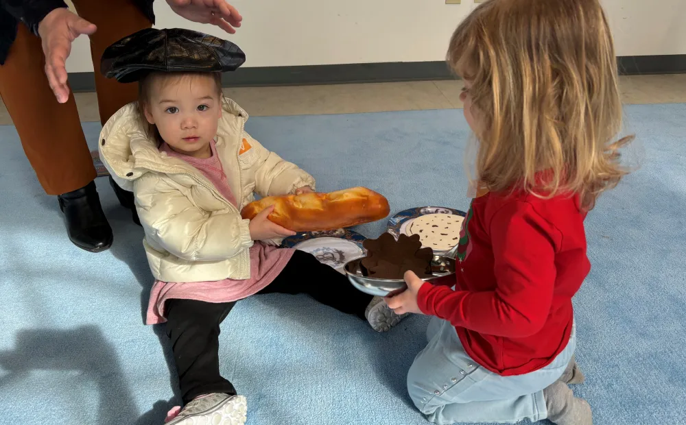 two preschoolers playing with food toys; one holds a baguette and wears a beret