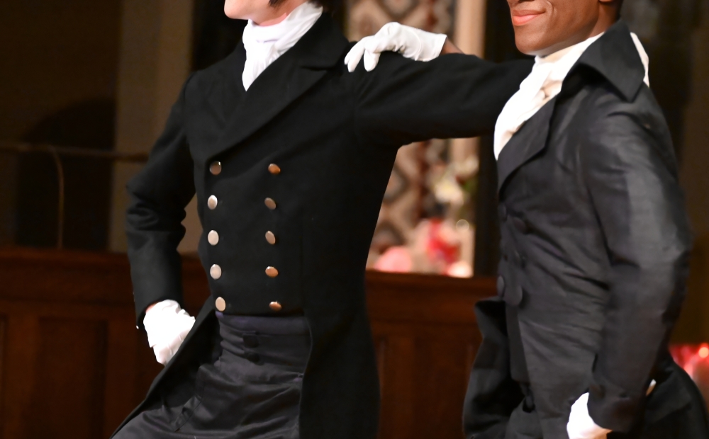 Image of two dancers, both wearing suits, posing with their arms around each other's shoulders. 