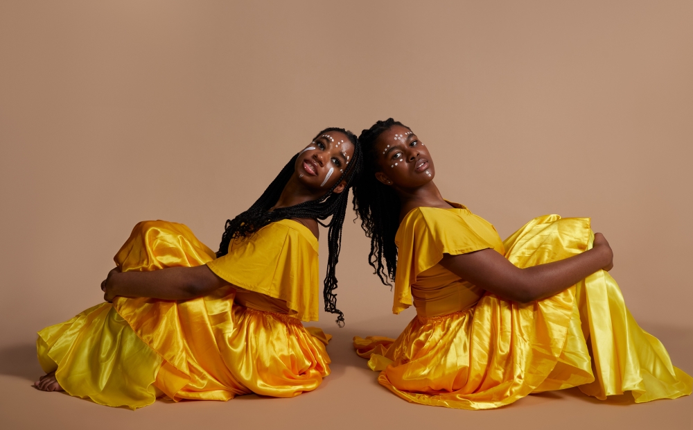 Image of two young dancers sitting on the ground, both in matching yellow costumes. They are both looking at the camera. 